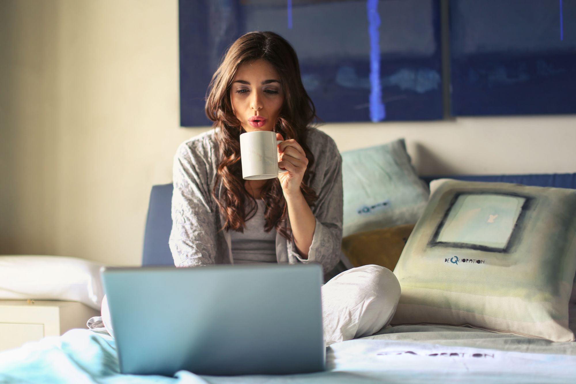 Woman drinking from a mug while using a laptop on a sofa