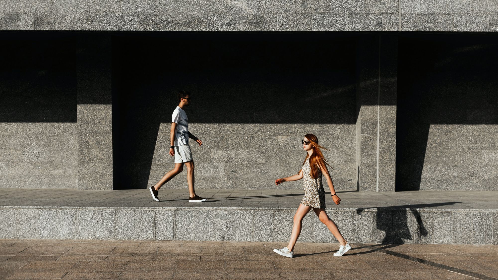 Two people walking in opposite directions beside a stone building